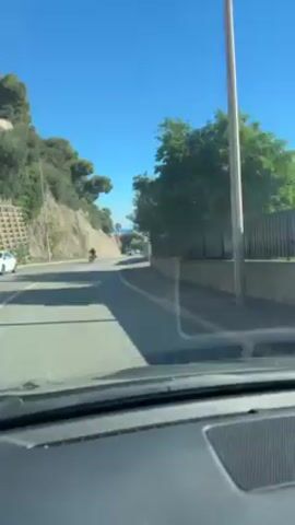 The video shows a car driving on a road towards the sea.  The sky is clear and blue. On the left of the road is a hillside with trees, and on the right is a fence and some buildings.  Another vehicle, possibly a scooter, is ahead on the road.  Several other cars are visible in the distance. The camera is positioned inside the car, looking out through the windshield.