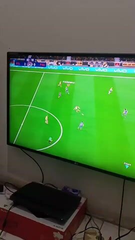 A television screen displays a soccer video game. The game is set on a green field with white markings. Several cartoon-like figures, representing soccer players, are scattered across the field. On the top of the screen, there is a scoreboard indicating the score is 2-0. To the left of the television, a black gaming console is visible, placed on top of a red and white box. Various cables are connected to the console and other devices. The overall environment appears to be a room with a plain wall.