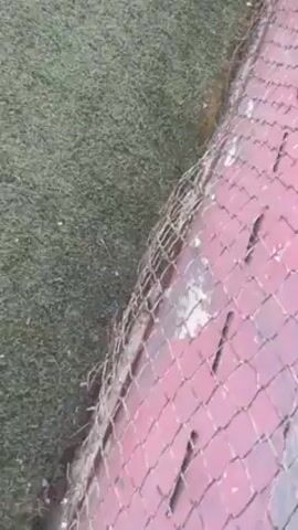 The video shows a basketball court seen through a chain-link fence. Several young men are playing basketball on the court. They are wearing various athletic clothes, including black shirts, white shirts, and navy blue shorts. The court's surface is painted with a combination of green, white, and red markings. The background includes trees and other greenery. The camera is positioned outside the fence, looking in towards the court.