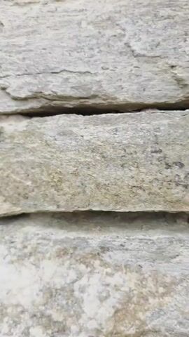 The video opens with a close-up view of stacked rough-hewn gray stones, likely part of a wall. The camera then pans right, revealing a cobblestone path winding through a rustic village. To the left of the path, more stone walls and buildings are visible, with traditional white houses featuring dark-trimmed windows. To the right, a rough stone wall continues to dominate the foreground. Further down the path, a large wooden gate is centered between two buildings. The scene is outdoors, with a cloudy sky and hints of greenery, suggesting an overcast day in a historic village setting.