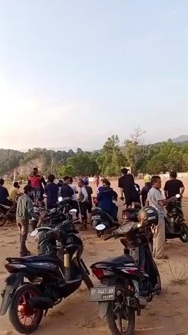 The video shows a crowded outdoor scene with many motorcycles parked. A group of people, mostly men, are gathered around the motorcycles, some standing and some sitting on the bikes. The ground appears to be dirt or sand, and there are hills with trees in the background. The sky is clear and bright, suggesting it is daytime. The lighting indicates it might be late afternoon or early evening. There are a few flags visible, possibly indicating a public gathering or event. The overall atmosphere appears casual, with people milling about and interacting.