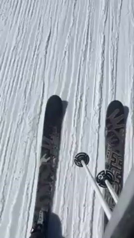 The video shows a first-person perspective of someone skiing down a groomed ski slope on a sunny day. The skis are visible at the bottom of the frame, and ski poles are also present. Other skiers are seen further down the slope. The surrounding environment includes snow-covered mountains and trees under a clear blue sky.