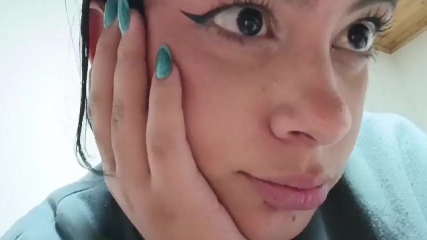 A close-up video captures a young woman's face in an indoor setting in Funza, Colombia. She has dark eyes, dark hair, and her skin shows small marks or freckles. She is wearing black winged eyeliner and a faint lip color. Her right hand, adorned with teal nail polish, rests against her cheek, supporting her head, as she looks directly at the camera. Her expression subtly shifts from a slight pout at the beginning to a neutral, listening look, before a soft, subtle smile appears on her lips towards the end of the clip. The background is out of focus, appearing as a light-colored wall. A male voice can be heard off-camera saying "Sí, con la WhatsApp," suggesting a conversation is taking place. No visible text is present in the video.