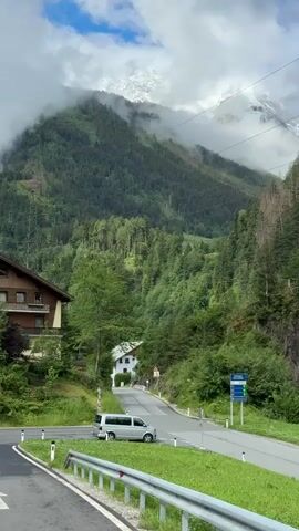 The video shows a scenic view of a mountain valley. A silver van is driving on a paved road that curves through the valley. On the left side of the road, there is a building with a brown facade and wooden balconies. Further up the road, a white house is visible. The valley is covered in dense green trees, and in the background, a snow-capped mountain peak is partially obscured by clouds. The sky is partly cloudy with patches of blue. There are power lines visible in the upper part of the frame. The overall environment appears to be a rural mountainous area.