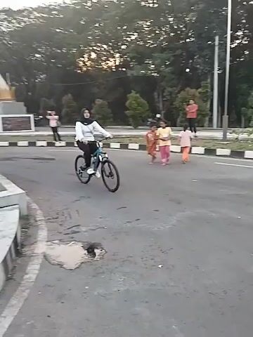 The video opens with a person riding a bicycle down a paved road. They are wearing a white long-sleeved shirt and black pants with a white head covering. They ride past several children on the side of the road and a large, multi-pronged statue in the background. The video then cuts to a close-up of a large statue of a muscular, ape-like figure holding a weapon. In front of the statue, there is a fountain with multiple jets of water shooting upwards and lights at the base of the jets. A man and a child are near the fountain. The video then shows another child sitting near a large, white letter "N" that is part of a larger sign. A flag is visible on a pole in the background.