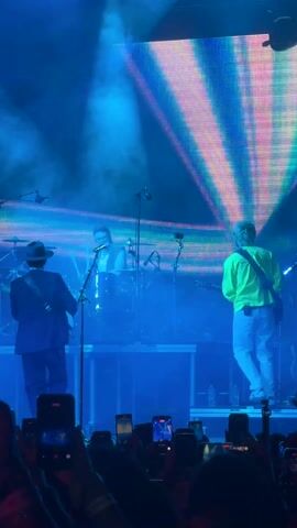 A live band plays on a stage under bright lights, a large LED screen behind them displaying colorful patterns. The band includes a drummer, who is front and center, wearing a white shirt and a vest. To his left, a guitarist in a dark suit plays, and to his right, another guitarist wearing a bright yellow shirt plays. The stage is slightly elevated, with a minimalist design consisting of clear stands. In the foreground, a crowd of people holds up their phones, recording the performance. The lighting changes, casting hues of blue, pink, and green onto the stage and the musicians.