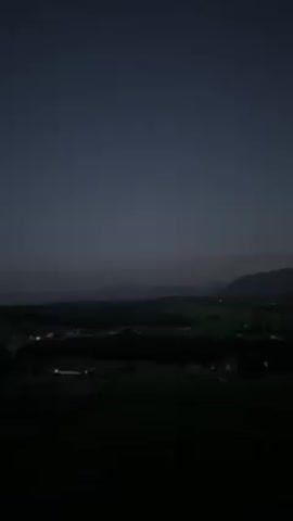 The video shows a night landscape with a full moon rising over distant mountains. The foreground features dark fields and hints of small settlements with lights. The sky is a deep blue, gradually lightening towards the horizon. There are no people visible in the frame.