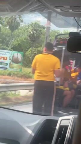 This media file shows a scene from a vehicle, likely a car, looking through the windshield and side window. The main focus is on a tuk-tuk, a common form of motorized rickshaw in Thailand.

**Subjects:**
Several people are visible inside and around the tuk-tuk. In the foreground, a person wearing a bright yellow t-shirt and black shorts stands, their back to the camera. To their right, another person in a purple t-shirt with white text is partially visible. Inside the tuk-tuk, multiple individuals are seated, some wearing colorful clothing. One person in the tuk-tuk wears a yellow shirt and dark shorts, while others are obscured by shadows or clothing. The tuk-tuk itself is a black, open-air vehicle with a roof. In the background, trees and foliage suggest an outdoor, possibly tropical setting.

**Activity or Event:**
The scene appears to capture people in or around a tuk-tuk, possibly in transit or waiting. The tuk-tuk is likely being used for transportation, a common sight in Tha Sala, Thailand.

**Notable Details:**
The lighting suggests it is daytime, with bright sunlight. The presence of lush greenery points to a warm climate. The image is somewhat blurry, suggesting it was taken while moving or from a distance. The people appear to be casually dressed, fitting for a warm environment. There are no strong emotional cues discernible due to the quality and framing of the image.

**Visible Text:**
On the purple t-shirt of the person on the right, there is white text that is partially visible and difficult to read clearly. Some characters appear to be Thai script.