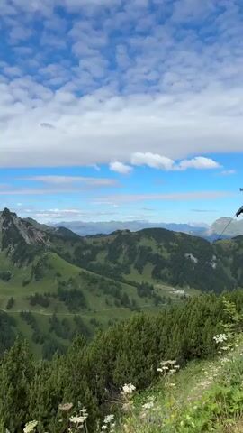 The video pans across a scenic mountain landscape on a partly cloudy day. The foreground features lush green pine trees and some wildflowers. Beyond the trees, rolling green hills covered in vegetation and scattered with trees dominate the middle ground. Winding roads are visible on the hillsides. In the background, rugged, rocky mountain peaks rise up against a bright blue sky with scattered white clouds. Small clusters of buildings, resembling a village, can be seen nestled in the valleys between the mountains. The camera movement is slow and steady, offering a panoramic view of the natural environment. There are no people or animals visible in the video.