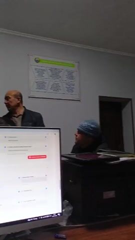 The video shows a room with three men working at desks with computers. The men are dressed in casual clothing and wearing beanies. They appear to be filling out an online form on the computer. The form appears to be in Uzbek and has fields for information about a respondent. There are Uzbek flags visible on the desks. The room is decorated with posters and certificates. The walls are light grey. The lighting is somewhat dim.