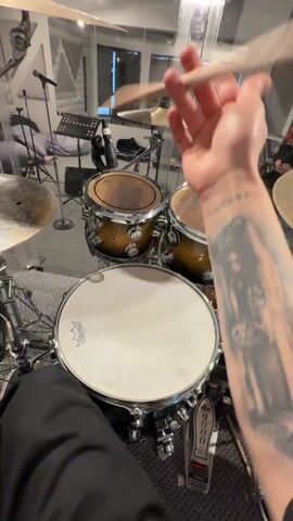 From a first-person perspective, a person is holding a drumstick and performing a drumstick twirl in front of a drum set. The drum set is in a soundproofed room. The drum set has two toms, a snare drum, a bass drum and cymbals. The drummer has a tattoo on their forearm. There is a music stand and microphone visible in the background of the soundproofed room.