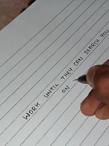 A person's hand is writing on lined paper. The writing says "WORK UNTIL THEY CAN SEARCH YOU ON 'GOOGLE'". The paper is white and lined with thin gray lines. The pen used is black. The background is out of focus and appears to be a textured surface, possibly wood or fabric.