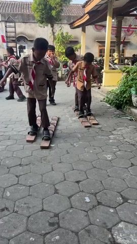 Four young boys wearing scout uniforms are participating in a game of synchronized walking on wooden planks. The planks are attached to their feet, and each boy has two planks, one for each foot, positioned side-by-side. They are walking in a line, attempting to move in unison. The boy in the front left is slightly ahead of the others. The boy behind him is closely following. Two other boys are further back, also walking on their planks. They are on a paved area with hexagonal tiles. In the background, there is a building with a tiled roof and some plants. There are also what appear to be motorcycles parked. The scene suggests an outdoor activity or event, possibly at a school or community center.