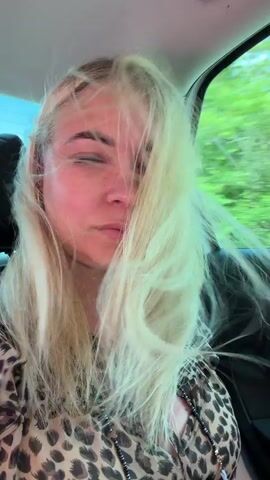 A blonde woman in a leopard print top is seated in the back of a moving car, with her left side facing the window. Initially, her long blonde hair is blowing across her face due to wind from an open window. She briefly raises her right hand to push some hair back from her face. She then settles back, resting her head on her left hand, propped by her left elbow, and gazes out the window with a calm expression as green trees and a blue sky blur past.