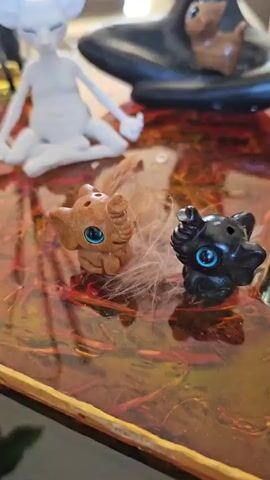 The video opens with a close-up on a decorative tabletop featuring two small, stylized animal figurines. The figurine on the left is light brown with blue eyes, and the one on the right is dark brown/black with blue eyes; both have wisps of white material around them. The table surface is reflective with an amber and reddish-brown swirling pattern. As the camera slowly pans upward, a white figurine resembling a large-eared creature or alien, sitting in a meditative pose, becomes visible in the center. To its left, a clear glass diffuser bottle with several black reeds is present. To the right of the white figurine, a black, stylized female figurine in a seated meditative pose with raised arms is visible, with a small brown animal figurine positioned near its base. The background shows a soft-focus beige sofa and a window, indicating an indoor setting.