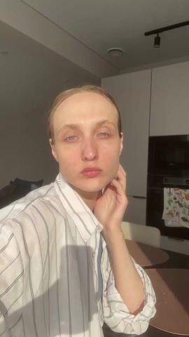 The video features a woman indoors in what appears to be a kitchen or dining area, potentially at home, as indicated by the background elements. She is Caucasian with fair skin, blonde hair pulled back, and blue eyes. She wears a light-colored, striped button-down shirt.

The woman has her hand gently touching her cheek/jawline on her left side. She looks directly at the camera for a moment, then looks slightly to her left, and then returns her gaze to the camera.

The environment is well-lit with natural sunlight, casting shadows across the scene. In the background, there is kitchen cabinetry, an oven, and a portion of a dining table. Overall, the scene is simple and focuses on the woman and her expressions.
