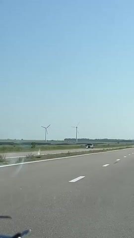 The video shows a view from inside a vehicle traveling on a highway. On the left side of the road, there are two wind turbines in a field. A dark-colored car is visible on the highway ahead. The sky is clear and blue, and the sun is shining. The scene appears to be a rural landscape.