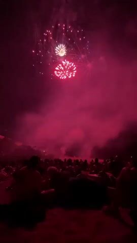 This is a vertical video of a fireworks display at night. In the foreground, a crowd of people is silhouetted against the dark sand and the illuminated sky. Their backs are to the camera as they watch the fireworks. In the sky, fireworks are exploding in bursts of red, pink, and green. Smoke from the fireworks drifts across the scene. The lighting is dim and atmospheric, with the bright bursts of light from the fireworks creating dramatic illumination. The video appears to be shot on a beach.