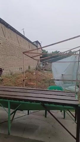 The media file shows an outdoor scene with a wooden table and benches in the foreground. Behind the table, there is a metal frame structure that appears to be a canopy or a shelter. Further in the background, a brick wall runs along the left side of the frame, and a greenhouse-like structure covered with plastic sheeting is visible on the right. The ground is covered with dry grass and debris. 

The setting appears to be a rural or semi-urban area, possibly a backyard or an open yard. There are no people visible in the media file. The objects in the frame include the table and benches, the metal frame, the brick wall, the plastic-covered structure, and some dry vegetation. There is also a hint of a baby stroller on the far left.

The weather appears to be overcast, suggesting it might be early morning, late afternoon, or a cloudy day. There are no explicit signs indicating the time of day or specific weather conditions beyond the general overcast sky.

The location cues, such as the brick wall and the style of construction, combined with the provided location context of Tarnau, Uzbekistan, suggest the scene is in Uzbekistan. The overall mood of the scene is quiet and somewhat desolate, possibly indicating abandonment or a work in progress. There is no visible text in the media file.