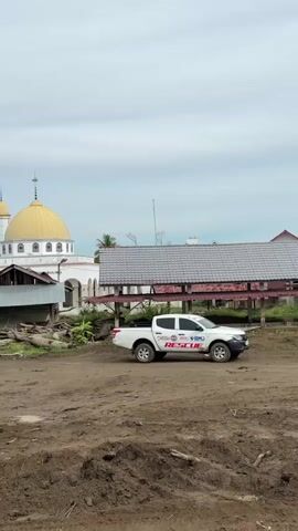 The video shows a wide shot of a rural or semi-rural area under an overcast sky. In the foreground, there is a large expanse of dirt and mud with some scattered debris, suggesting a recent disturbance or natural event. In the middle ground, a white pickup truck with "RESCUE" written on its side is parked on the dirt. To the left of the truck, a white mosque with a prominent golden dome is visible. Adjacent to the mosque, there are some damaged structures and debris. Further into the background, a series of buildings, some appearing damaged and others under construction or incomplete, are spread across the landscape. Palm trees and other vegetation are scattered throughout the scene. The overall impression is one of a community that has experienced some form of disaster or significant disruption, with rescue efforts potentially underway, as indicated by the presence of the rescue vehicle.
