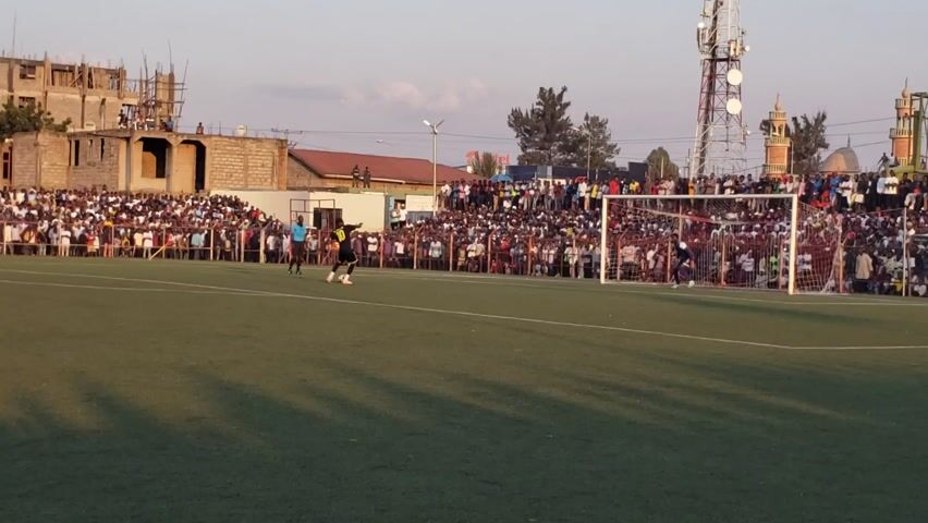 Here is a concise description of the video:

The video shows a soccer game on a green field, with large crowds of spectators watching from behind a perimeter fence. A player in a yellow and black uniform approaches the goal while the goalie is kneeling in front of the goal. A referee in a blue shirt stands in the middle of the field, facing the action. A building under construction and a cell tower are visible in the background. The sky is clear and the scene appears to be taking place during the day.