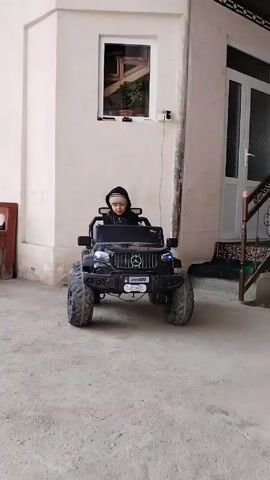A young child dressed in black, including a hooded jacket and beanie-style hat, is driving a black electric ride-on toy car, designed to resemble a Mercedes-Benz G-Wagon, on a concrete surface in what appears to be a covered patio or carport. The child drives forward with a quick burst of speed. The car has large, knobby tires and a faux license plate that reads "008." The background includes a white building with a small window and a door with white trim and glass panels. There are also some decorative iron railings visible and a glimpse of what looks like an indoor space with a patterned ceiling. A charging cable is plugged into the wall near the window. A wooden item and mat sit beside the wall.