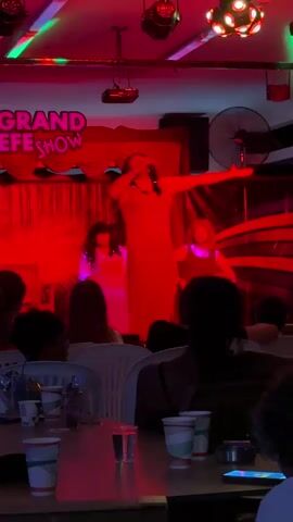 The video shows a drag queen performing on stage in front of an audience. The performer is wearing a long, flowing orange dress and is holding a microphone, singing and gesturing. The stage is lit with colorful lights, primarily purple, blue, green, and red, creating a vibrant atmosphere. In the background, there are curtains and a sign that reads "GRAND SHOW". There are other performers on stage, also dressed in elaborate attire. The audience is seated at tables in a dimly lit room, with cups and other items on the tables. The camera angle is from the audience's perspective, looking up at the stage. The video appears to be a recording of a live performance.