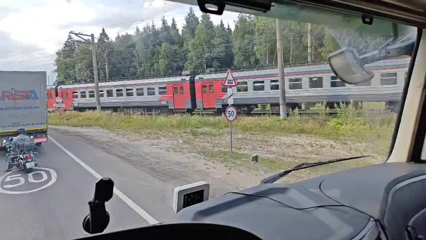 The video is shot from the perspective of a driver inside a vehicle, likely a truck, looking out through the windshield. In the foreground, the dashboard and part of the steering wheel are visible. A small electronic device is mounted on the dashboard. Outside, a red and white train is passing by on a railway track. To the left of the train, a road is visible with a motorcycle on it. There are traffic signs on the side of the road, including a speed limit sign indicating 50. The environment appears to be rural, with trees lining the background. The sky is overcast. The train is moving from left to right across the frame.