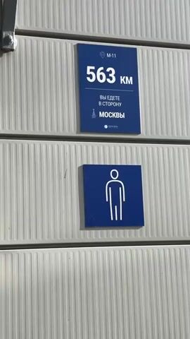 The video shows a white ridged wall with two blue signs. The top sign reads "M-11 563 KM YOU ARE GOING TOWARDS MOSCOW". The bottom sign is a restroom symbol. Below the signs is a dark gray door. Above the door is a white circular light fixture with a bird's nest on top of it. The nest appears to be made of twigs and mud. There are small dark specks on the light fixture, likely bird droppings. The wall and ceiling are illuminated by sunlight casting shadows.