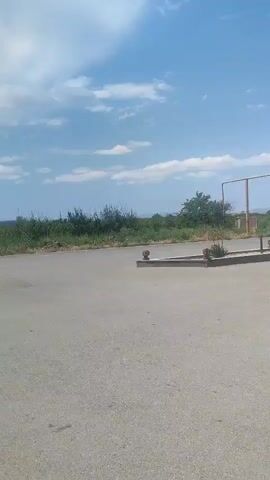 The video shows an exterior shot of an asphalt surface, possibly a road or parking lot, under a blue sky with scattered white clouds. In the background, there is a line of green vegetation and trees, with some distant hills or mountains visible. A yellow horizontal bar, possibly part of a structure or fence, is present in the middle ground. Several small, reddish-brown objects are lined up along a raised concrete barrier in the foreground. The camera pans slowly from right to left, showing different sections of the asphalt and the surrounding landscape. There are no people or animals visible in the frame. The lighting suggests it is daytime, likely a sunny day.