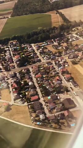 An aerial view shows a suburban residential area with houses, streets, and some agricultural fields in the background. The houses are mostly single-family dwellings with red-tiled roofs. Many properties have swimming pools visible. The streets are paved and wind through the neighborhood. Some areas have trees and vegetation. The overall impression is that of a peaceful, rural or semi-rural community.