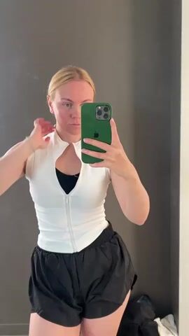 A blonde woman with her hair pulled back is standing in front of a dark gray wall, taking a selfie with her green-cased smartphone. She is wearing a white, sleeveless, zip-up top over a black sports bra and black shorts. Her left hand is on her hip, and her right hand is holding the phone. She is looking at the phone screen, with her lips pursed slightly. There is a glimpse of a doorframe to her left and a black bag and some clothing to her right. The lighting is even, and the focus is on the woman.