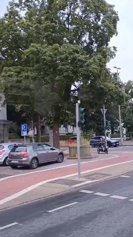 The video pans across a street scene. In the foreground, a road with dashed white lines marking lanes is visible. A red and white striped bike lane runs alongside. A grey car is parked on the left side of the road, and a person rides a three-wheeled vehicle in the bike lane. Traffic lights are visible, one of which is red. Trees with green foliage line the street. In the background, a building with white walls and a few cars can be seen. The sky is overcast.