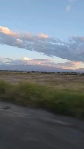 The video shows a view from a moving vehicle, likely a car, of a vast field of dry grass with patches of green vegetation. In the distance, there are snow-capped mountains, partially obscured by clouds. The sky is blue with scattered white and gray clouds, some tinted pink by the sun. The road is visible in the foreground, and the camera is panning from left to right, following the movement of the vehicle. There are some dark indistinct structures in the mid-ground.