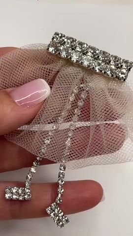 A hand with a French manicure is holding a delicate hair accessory. The accessory is made of sheer, blush-colored mesh fabric that is gathered at the top. Attached to the mesh is a rectangular bar embellished with two rows of sparkling rhinestones. Dangling from the mesh are two strands of rhinestones, each ending in a cluster of rhinestones. The background is a plain, white surface.