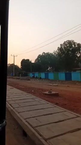 The video shows a view from inside a yellow motorized tricycle, often called a tuk-tuk. The driver, whose hands are visible, is driving down a road with other tuk-tuks, cars, and pedestrians. The road appears to be under construction, with patches of dirt and concrete dividers along the side. A blue and green painted wall lines part of the road. The sky is a light, pale color, indicating early morning or late evening. The driver honks the horn multiple times.