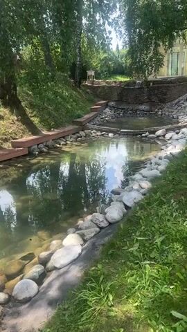 A video captures a serene natural landscape with a man-made water feature. A gently flowing stream, bordered by large, smooth river stones, meanders through lush green grass and mature trees. The water is clear, reflecting the dappled sunlight and the surrounding foliage. Wooden planks are laid out as a pathway along one side of the stream, leading towards a small, stone-lined pond or pool.

In the background, a tiered structure made of stone creates a subtle waterfall effect, feeding into the pond. Beyond this, a building with large windows and a pale exterior is visible, suggesting a residential or recreational setting. A white chair sits on a grassy mound near the building, and further back, more trees and vegetation create a sense of depth. The overall atmosphere is tranquil and inviting, hinting at a warm, sunny day in a rural or semi-rural location. The scene appears to be set in Dal'nie Prudishchi, Russia. No people are actively visible in the frame, and there is no visible text. The time of day appears to be daytime, with bright sunlight indicating fair weather. The focus is on the natural beauty and the integration of the water feature into the landscape.