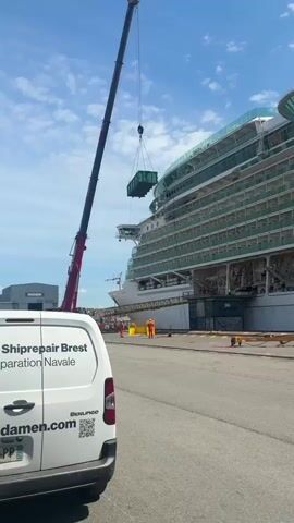 Here's a description of the video:

The video shows a large cruise ship docked at what appears to be a port or shipyard. A tall, red-and-black crane is lifting a green container high in the air, presumably to load it onto the ship or move it to another location on the dock. Several workers in orange jumpsuits are visible around the base of the ship.  

A white van with the text "Reprepair Brest" and "Construction Navale" is parked in the foreground, partially obstructing the view. The sky is blue with some clouds. The environment appears to be a busy industrial area.