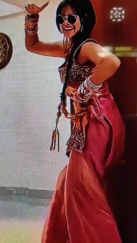 The video captures a young woman dancing in a room. She is wearing a pink and gold traditional Indian outfit, complete with bangles and earrings. She is also wearing sunglasses and headphones. The woman is in motion, with her arms raised and her body swaying. The background of the video is blurred, but it appears to be a domestic setting with some furniture visible. The lighting in the room is warm and inviting, and the overall atmosphere of the video is one of joy and celebration.