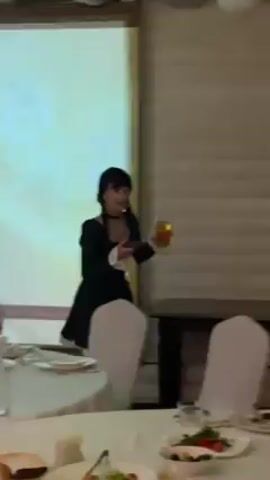 A woman dressed as Wednesday Addams stands behind a dining table holding a drink and her phone. She is wearing a black dress with white cuffs and a collar, black tights, and black pigtails. The dining table is set with white tablecloths and chairs. In the background, there is a window and a white cabinet. The overall setting appears to be a party or reception.