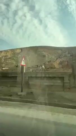The video, shot from a moving vehicle, depicts a roadside scene. On the right side of the road, there's a weathered concrete wall with remnants of damaged or unfinished structures. Behind the wall, a dry, hilly landscape extends towards the horizon, beneath a sky with scattered clouds. The foreground shows the asphalt road, appearing slightly dirty. Some sparse vegetation and debris are visible behind the wall. The overall impression is of an arid, possibly neglected area.