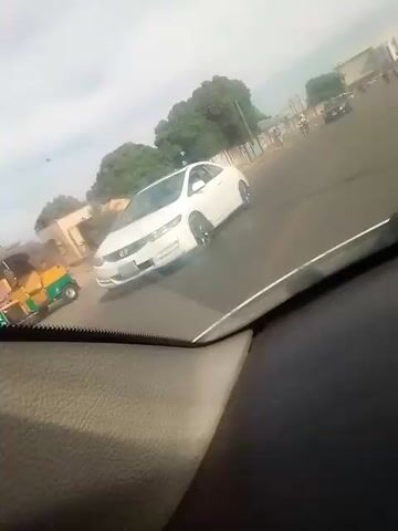 The view is from inside a vehicle traveling on an asphalt road. A red Honda Accord passes in the opposite direction. There is a concrete wall alongside the road with painted warnings such as "NO PARKING" and "DO NOT URINATE HERE." A person is walking on the side of the road next to the wall. Trees and other vehicles are visible in the distance. The sky is overcast.