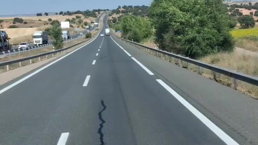 The video captures a view from a vehicle traveling on a highway. The road has multiple lanes, marked with white lines. Guardrails are visible on both sides of the road. The landscape is hilly and covered with vegetation, including trees and dry grass. In the distance, there are other vehicles, including trucks and cars, and a billboard. The sky is clear and the sun is shining, suggesting it is daytime. The video shows the road ahead curving and ascending into the distance.