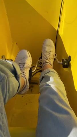 A first-person perspective shot shows a person's feet in white sneakers and blue jeans inside a bright yellow pedal boat. The camera pans to show other yellow pedal boats on a calm lake, some with people in them, and a shoreline with trees and people standing near the water.