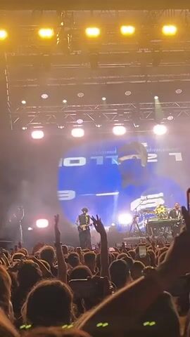 A large crowd of people are at a concert. A man is playing a guitar on stage, while another person plays keyboards. A large screen behind the performers displays a picture of a man's face. The stage is lit with bright lights.