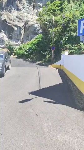 A paved road curves uphill, flanked by a rocky cliff face and lush green vegetation. On the left side of the road, two cars are parked, one silver and one dark gray. A white building with green shutters is visible behind the cars. A street sign with a blue "P" is mounted on a pole on the right side of the road. A red and white circular traffic sign is also present. The sky is clear and blue.