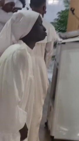 The video shows a group of people, likely men and women, dressed in white robes and head coverings. They appear to be in a place of worship or a formal gathering. The person in the foreground, a Black individual with dark skin, is wearing a white robe and a white head covering. Their head is turned to the right, and their mouth is open, suggesting they are speaking or singing. Their hands are raised, with fingers spread, in what could be a gesture of prayer or praise. Other individuals in white robes are visible in the background, some looking towards the foreground person and others looking away. The environment appears to be indoors, with white walls and a tiled floor. There is a green plant visible to the right of the frame. The lighting is bright, indicating artificial light or good natural light. The overall atmosphere seems solemn and devotional.