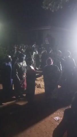 The video shows a gathering of people outside at night. They appear to be dancing or moving to music, though details are obscured by darkness and bright light. There's a light source from above. The ground seems to be dirt.