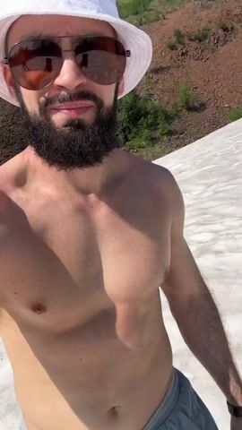 A shirtless man with a beard, sunglasses, and a white bucket hat takes a selfie. He is wearing gray shorts with a drawstring. The man is standing in a snowy or icy area with a rocky, wooded hillside in the background. The sky is clear and blue. The man smiles at the camera.