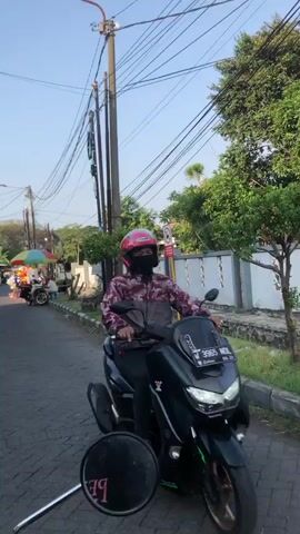 A person wearing a red helmet, a camouflage jacket, and a black face mask rides a dark green scooter on a paved road. The scooter has a license plate reading "B 3965 NOL." In the foreground, a circular mirror reflects the rider and the scooter. The background features utility poles with wires, trees, and a white wall. To the left, a street vendor with a colorful umbrella is visible, along with another scooter. The scene appears to be an outdoor street setting in daylight, possibly late afternoon given the warm lighting. The weather seems clear. The location is identified as Kludan, East Java. The overall mood is casual and everyday.